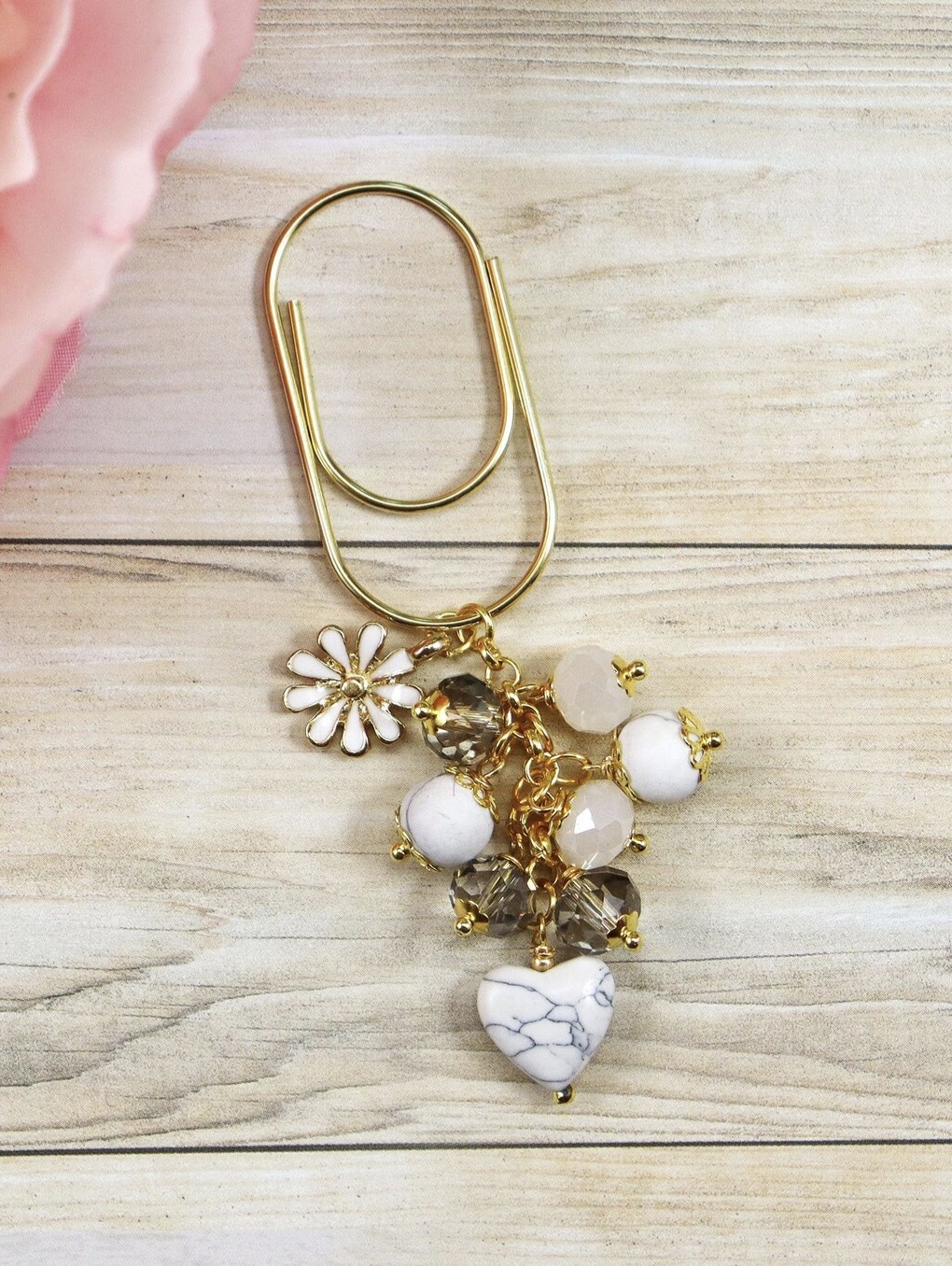 This image shows a gold toned oversized paperclip with a golden chain strong with white air descent beads, a heart shaped marble bead, and a daisy charm. This planner charm is sitting on top of a light colored wood background with sunflower petals showing in the corner.